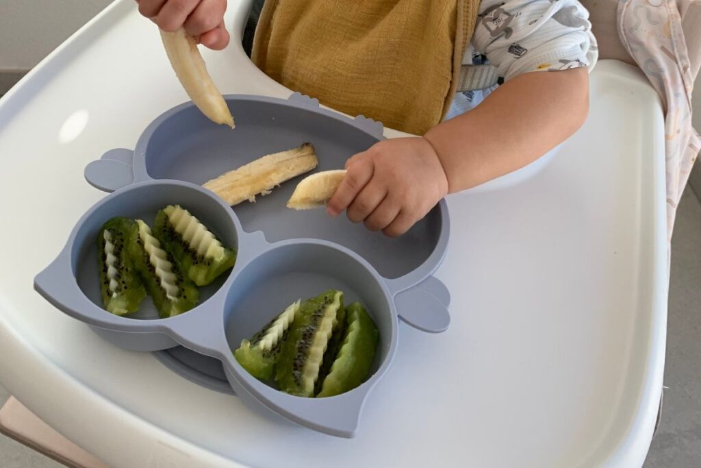Kinderhände greifen nach Beikost auf einem unterteilten Teller – Banane und Kiwi beikostgerecht angerichtet